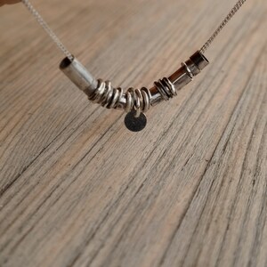 May include: A silver necklace with a pendant made of multiple silver rings and a small round disc. The necklace is on a wooden surface.