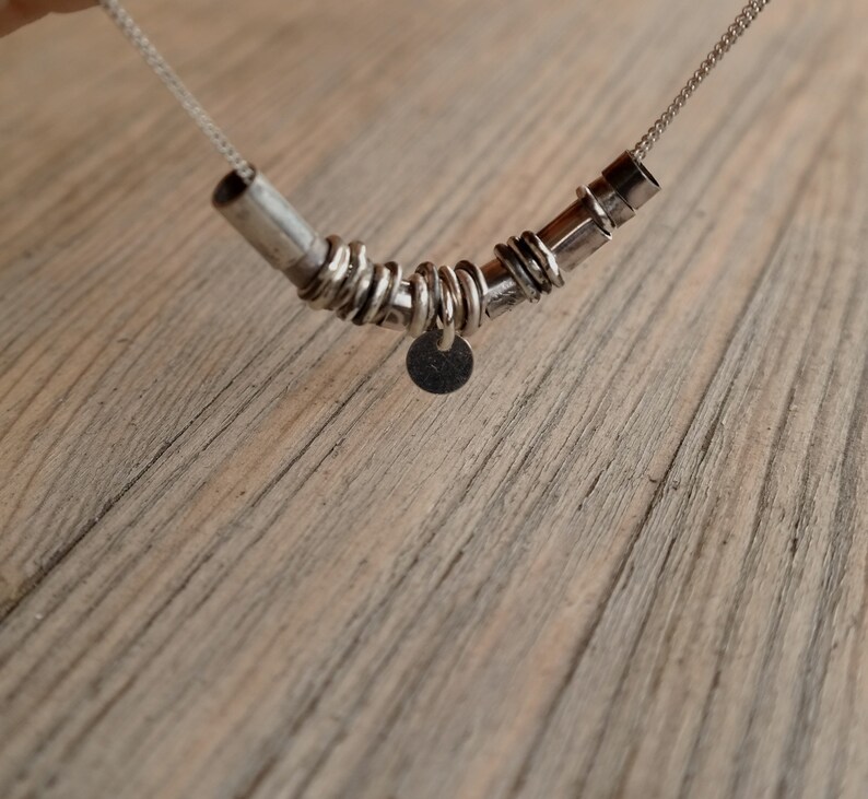 May include: A silver necklace with a pendant made of multiple silver rings and a small round disc. The necklace is on a wooden surface.