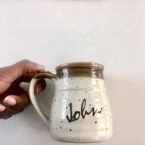 May include: A speckled ceramic mug with a brown rim and handle. The mug is off-white with black speckles and the name "John" written in black script. The mug is held by a hand.