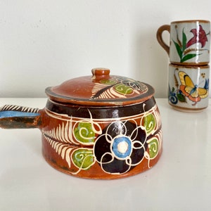 May include: A ceramic, lidded soup pot with a handle, featuring a brown and black floral design with white, green, and blue accents. The pot is accompanied by two decorative mugs with floral and butterfly motifs.