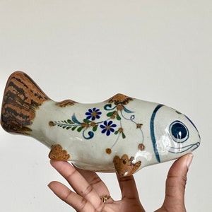May include: A ceramic fish figurine with a white body, brown tail and fins, and blue floral accents. The fish has a detailed design with a painted eye and mouth. The figurine is held in a hand.