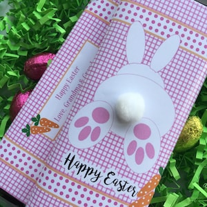 May include: Two chocolate bars wrapped in pink gingham paper with a white bunny design and the words "Happy Easter." The bars are decorated with a carrot and polka dot ribbon. The bars are surrounded by green Easter grass and chocolate eggs.