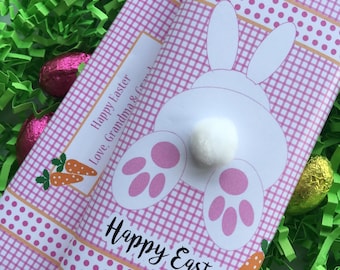 May include: Two chocolate bars wrapped in pink gingham paper with a white bunny design and the words "Happy Easter." The bars are decorated with a carrot and polka dot ribbon. The bars are surrounded by green Easter grass and chocolate eggs.