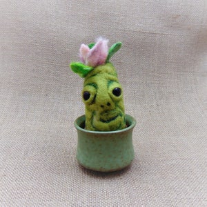 May include: A green felt plant with a pink flower and a face, sitting in a green ceramic pot.