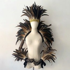 May include: A mannequin displays a feathered headdress and shoulder piece. The headpiece features a gold band. The feathers are brown, black, and tan. Black straps and wrist cuffs with black ribbons complete the ensemble. The overall theme is costume or theatrical.