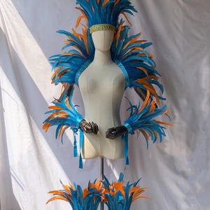 Könnte beinhalten: Eine Schaufensterpuppe präsentiert ein lebendiges Kostüm mit blauen und orangefarbenen Federn. Das Ensemble umfasst ein Kopfschmuck, Schulterstück, Armbänder und Beinmanschetten. Das Kostüm hat ein goldenes Band und blaue Bänder.