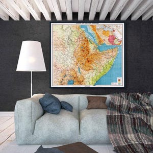 Puede incluir: Un mapa vintage enmarcado de África y territorios circundantes, con una representación detallada de la topografía y los límites políticos. El mapa está en un marco negro, sobre un sofá gris claro con cojines azules y marrones.