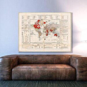 Op de afbeelding: Een ingelijste vintage wereldkaart met rode accenten, weergegeven boven een bruine leren bank. De kaart bevat gedetailleerde geografische informatie en is gemonteerd op een lichtgekleurde achtergrond. Een vloerlamp staat rechts.
