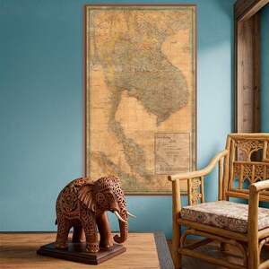 May include: A detailed, wooden elephant sculpture with intricate carvings stands on a dark wooden base. Behind it, a vintage map of Southeast Asia hangs on a blue wall, and a bamboo chair is to the right.