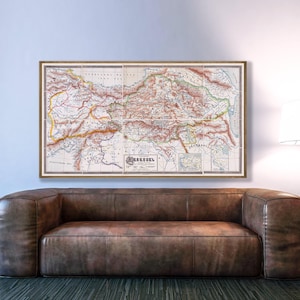 Puede incluir: Un mapa antiguo enmarcado que muestra una representación detallada de una región, con varios colores y líneas intrincadas. El mapa se exhibe sobre un sofá de cuero marrón, creando una estética vintage. El marco del mapa es de un tono cálido y neutro.