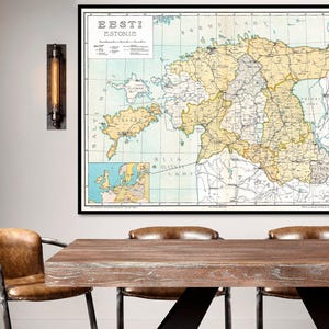May include: A vintage map of Estonia, titled 'Eesti Estonie' in black text. The map is printed on a light blue background with brown lines for roads and rivers. The map is framed in a black frame and hanging on a white wall. The map is surrounded by a wooden table with three brown leather chairs.