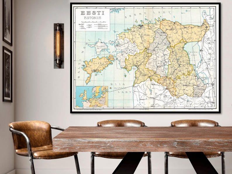 May include: A vintage map of Estonia, titled 'Eesti Estonie' in black text. The map is printed on a light blue background with brown lines for roads and rivers. The map is framed in a black frame and hanging on a white wall. The map is surrounded by a wooden table with three brown leather chairs.