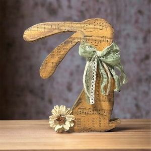 May include: A wooden bunny rabbit figurine decorated with musical notes and a green bow. The bunny is holding a white flower with a brown button in the center. The bunny is sitting on a wooden surface.