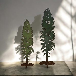 May include: Two wooden cutout trees, one larger than the other, painted in shades of green and brown. The trees are standing on a light brown surface.