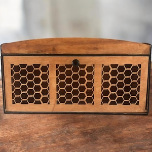 May include: Wooden chest with a light brown finish and a dark brown trim. The front features three panels with a honeycomb pattern. A small, round, black knob is centered on the top panel. The chest is sitting on a wooden surface.
