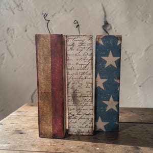May include: Three decorative wooden blocks with patriotic designs. One block features red and gold stripes, another has handwritten script, and the third displays a blue background with white stars. Each block has a wire accent.