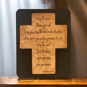 May include: A wooden cross with a black frame, featuring a bible verse from Numbers 6:24-26. The verse reads: "May the Lord bless you and keep you. May the Lord make His face shine upon you and be gracious to you. May the Lord turn His face upon you and give you peace."
