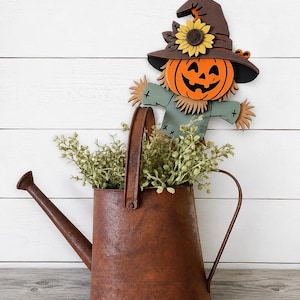 May include: A rustic, weathered brown metal watering can filled with light green faux foliage. A layered wooden cutout of a scarecrow with an orange pumpkin head, a brown witch hat adorned with a yellow sunflower, and a sage green shirt.