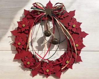 May include: A festive red poinsettia wreath with a star-shaped accent and small bells. The wreath features red paper flowers with yellow centers and is adorned with a raffia bow and ribbons. A decorative piece for the holiday season.