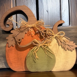 May include: Wooden pumpkin decoration in orange, green, and cream colors. Each pumpkin has a wooden stem and leaf accents, with raffia bows. The pumpkins are layered to create a 3D effect, perfect for fall decor.