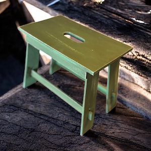 May include: A small, olive green wooden step stool with a rectangular top and a handhold cutout. The stool has four angled legs and a simple, sturdy design. The paint has a slightly distressed look.