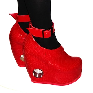Könnte beinhalten: Ein Paar rote Plateau-Keil-Schuhe mit Knöchelriemen. Die Schuhe haben eine strukturierte, lederartige Oberfläche und einen runden Ausschnitt an der Seite des Keils.