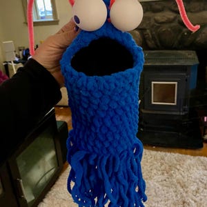 May include: A blue crocheted puppet with large white googly eyes and pink pipe cleaner antennae. The puppet has a large open mouth and a long, shaggy blue beard.