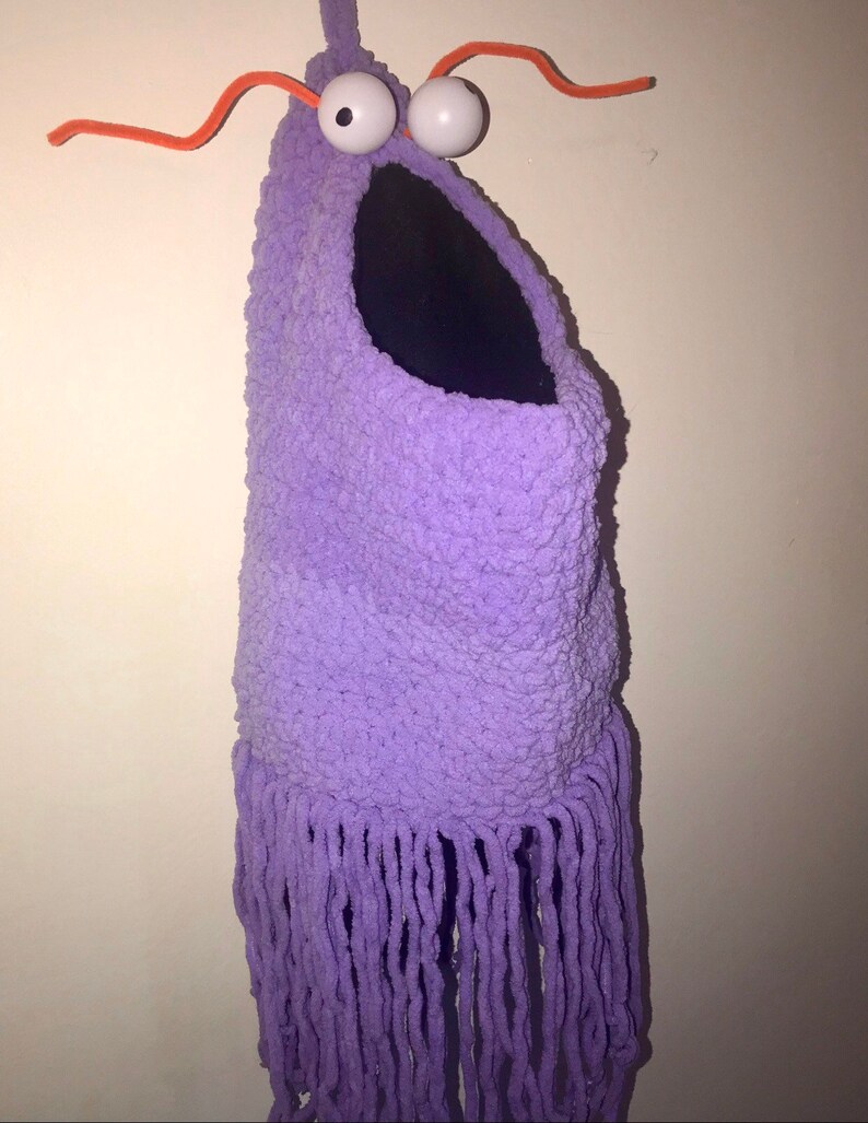 May include: A purple crocheted monster with a large mouth and googly eyes. The monster has long purple fringe hanging from the bottom.