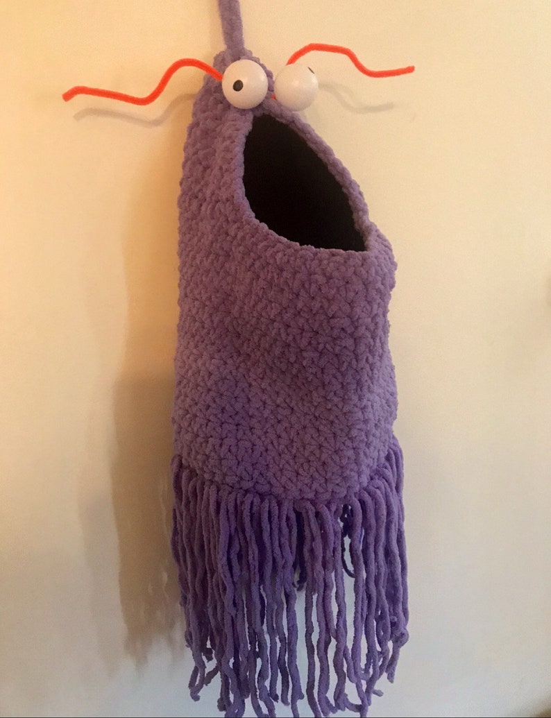 May include: Purple crocheted hanging toy with a large mouth and googly eyes. The toy has a fringe at the bottom and red pipe cleaner antennae.