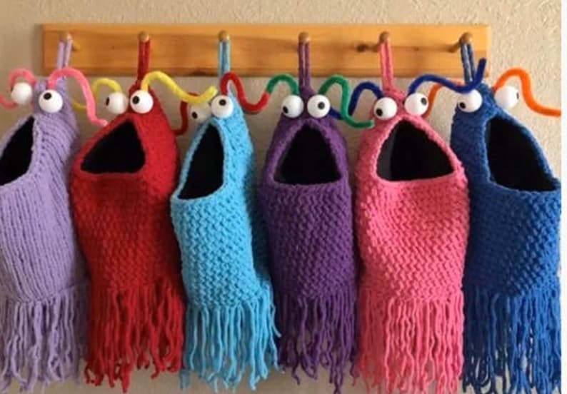 May include: Six colorful knitted monster headbands with googly eyes and yarn antennae. The headbands are hanging on hooks and have fringe at the bottom. The colors are purple, red, blue, purple, pink, and blue.