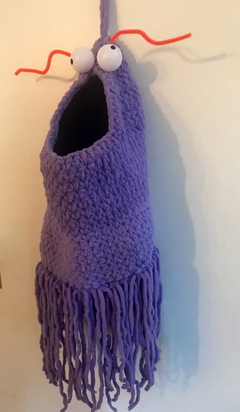 May include: Purple crocheted monster toy with googly eyes and orange pipe cleaner antennae. The monster has a large open mouth and a fringe of yarn around the bottom.