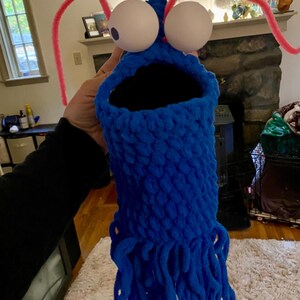 May include: A blue crocheted puppet with large white googly eyes and pink pipe cleaner antennae. The puppet has a large open mouth and a long fringe of blue yarn.