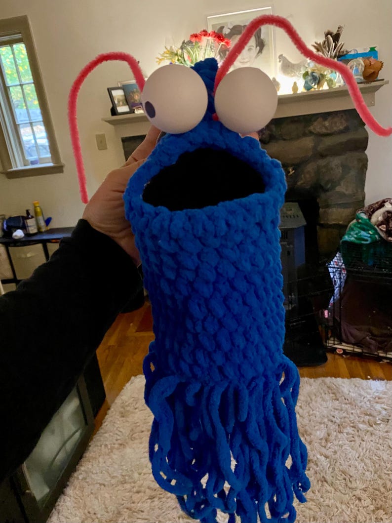 May include: A blue crocheted puppet with large white googly eyes and pink pipe cleaner antennae. The puppet has a large open mouth and a long fringe of blue yarn.