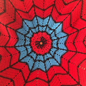 May include: A crocheted blanket with a red, blue, and black spiderweb design. The center of the blanket features a small red flower with a black center.