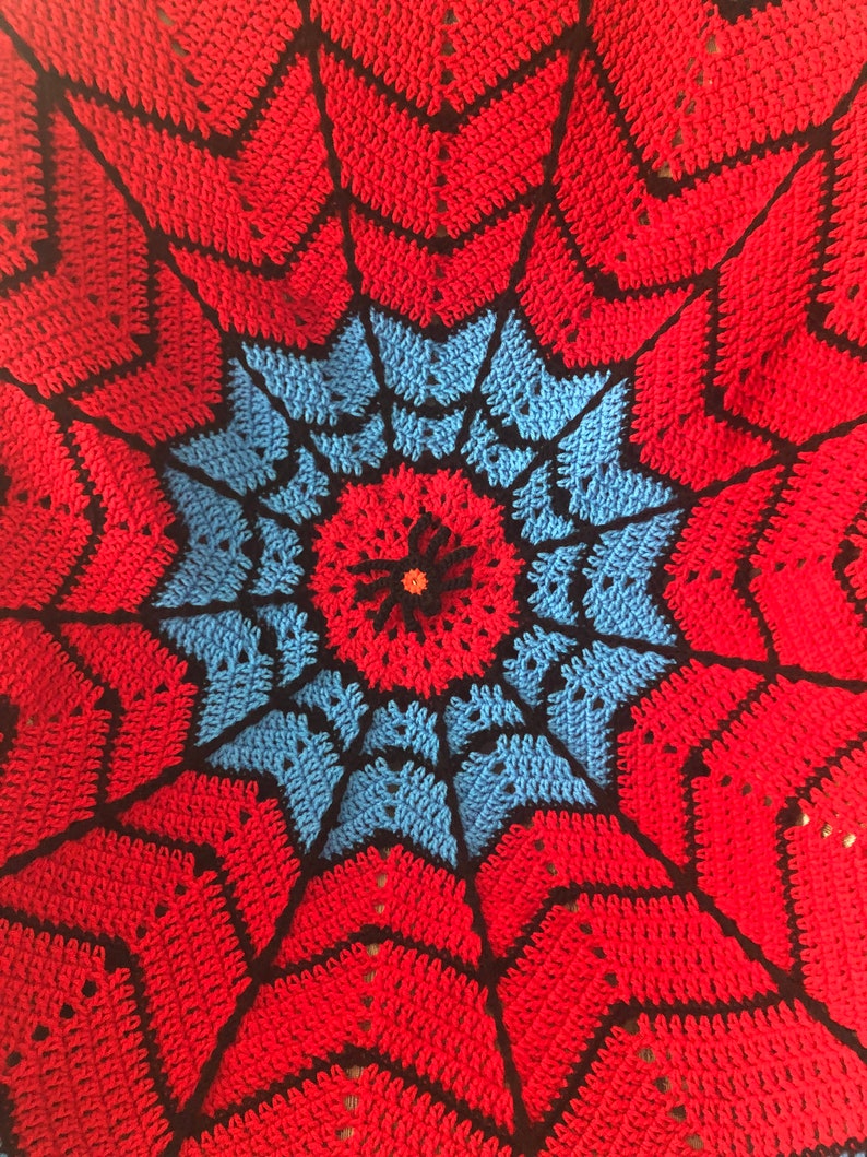 May include: A crocheted blanket with a red, blue, and black spiderweb design. The center of the blanket features a small red flower with a black center.