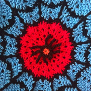 May include: A crocheted mandala with a red and black spider in the center. The mandala is made with blue, black, and red yarn.