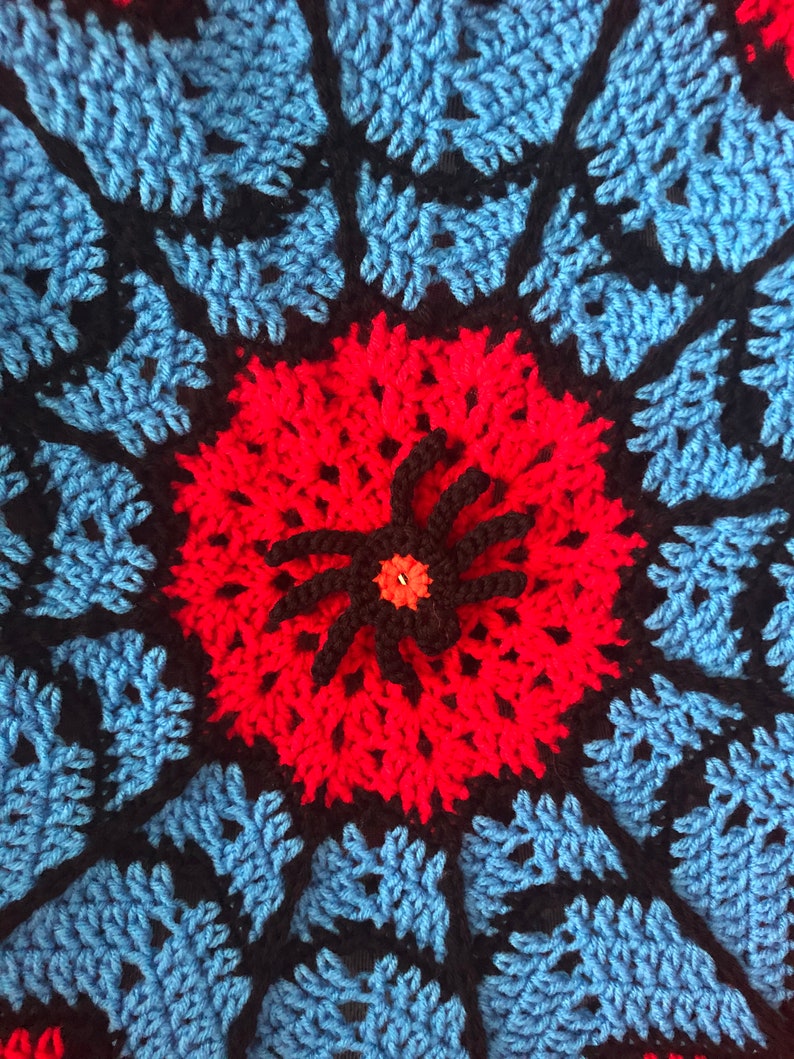 May include: A crocheted mandala with a red and black spider in the center. The mandala is made with blue, black, and red yarn.