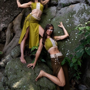 May include: Two-piece outfits consisting of a sleeveless olive green crop top with a central light-coloured panel and a matching high-waisted skirt featuring side lacing, a wide waistband, and a high leg slit, worn by two individuals in a natural setting.