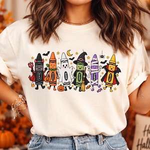 May include: A cream-coloured t-shirt featuring a graphic of six anthropomorphic crayons dressed in Halloween costumes. The crayons are labelled Black, Orange, White, Green, Purple, and Yellow, and are surrounded by small bats, stars, and a crescent moon.