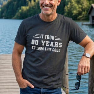 May include: A charcoal grey short-sleeved crew neck t-shirt featuring white text that reads IT TOOK 80 YEARS TO LOOK THIS GOOD, with horizontal lines flanking the top line of text, worn with blue denim jeans and a wristwatch.