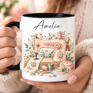 Pode incluir: Uma caneca de cerâmica branca com asa e rebordo pretos, com uma ilustração impressa de uma máquina de costura cor-de-rosa rodeada por padrões florais, carretéis de linha, tesouras, uma almofada de alfinetes e uma fita métrica, com o nome Amelia.