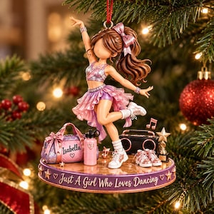 Könnte beinhalten: Ein dekoratives Ornament mit einer braunhaarigen Tänzerin in einem rosa-violetten Outfit auf einem runden Sockel mit der Aufschrift 'Just A Girl Who Loves Dancing'. Zubehör umfasst eine 'Isabella'-Sporttasche, eine Trinkflasche und einen Pokal.
