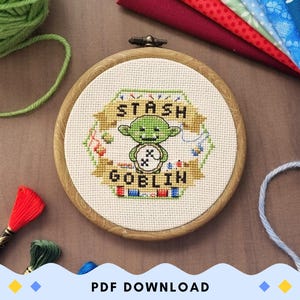 以下が含まれることがあります： 緑色のゴブリンが宝箱を持っており、「Stash Goblin」という文字が書かれたクロスステッチのパターン。パターンは木製の刺繍枠に表示されています。