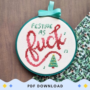 Könnte beinhalten: Ein runder Stickrahmen mit weißem Stoff, auf dem der Text "FESTIVE AS FUCK" in rotem und grünem Garn im Kreuzstich gestickt ist. Unter dem Text befinden sich ein kleiner grüner Weihnachtsbaum und Noten. Oben ist ein blaugrünes Band befestigt.