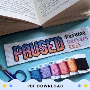 May include: A cross-stitched bookmark featuring the word "PAUSED" in a gradient design, with "Resume, Restart, Exit". The image includes DMC thread spools, scissors, and an open book. The bookmark is on a blue surface.