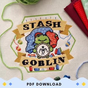 May include: Cross-stitch design featuring a green frog wearing a blue outfit and holding a clock. The design includes the words "STASH GOBLIN" on a banner, with sewing supplies and fabric in the background. A ball of green yarn and fabric are also visible.