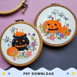May include: Two cross-stitch embroidery hoops with Halloween-themed designs. One features a black cauldron with an orange face and bubbling orange liquid. The other has an orange pumpkin with a carved face. Both have floral accents and are set in wooden hoops.