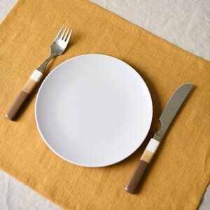 May include: A white ceramic plate with a silver fork and knife on a mustard yellow placemat.