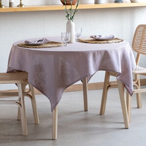 Puede incluir: Un mantel de lino de color lila claro cubre una mesa redonda de madera con dos sillas de mimbre. Un jarrón con flores blancas se coloca en el centro de la mesa, con dos cubiertos a cada lado.