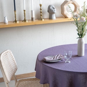 May include: A round, purple linen tablecloth covers a wooden table with a single woven chair. A glass of water and a folded napkin are on a white plate on the table.